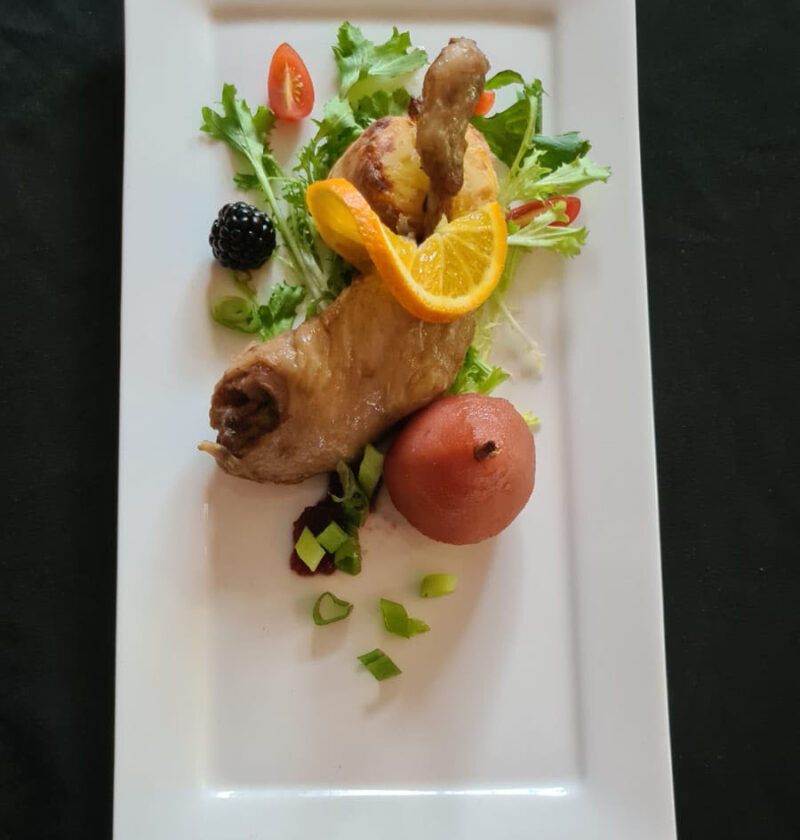 Wit rechthoekig bord op zwarte tafel met eendenbout, rode kool een stoofpeertje en aardappelgratinee prachtig opgemaakt met een schijfje sinaasappel.